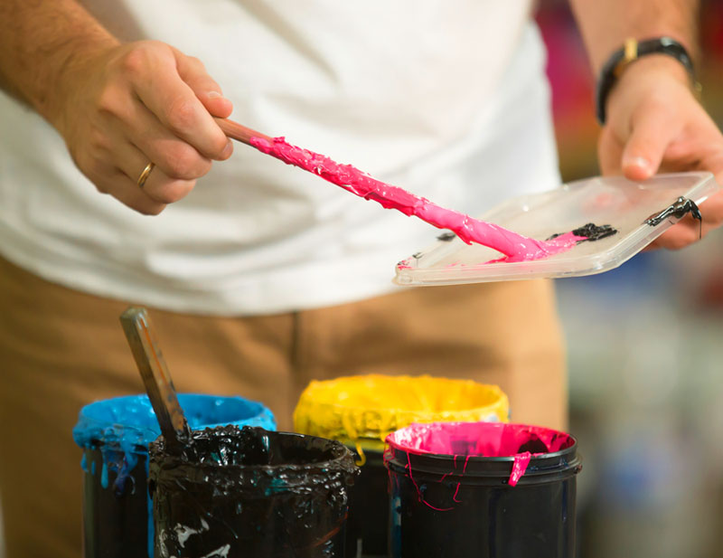 This photo shows a person holding a paintbrush with four colors of dye in front of them--pink, blue, black, and yellow--representing the screen-printing process.