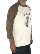 Angled view of man wearing a Kaoru Betto baseball shirt, with olive green raglan sleeves, a beige body, and a black screenprint of a Japanese baseball player